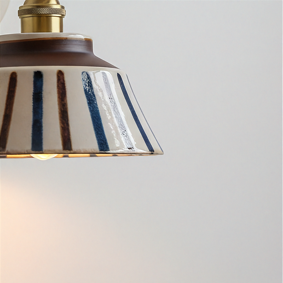 Close-up of a ceramic pendant light shade with vertical blue and brown stripes on a cream glaze, with a brass-toned metal fixture and warm glowing light against a light gray background.
