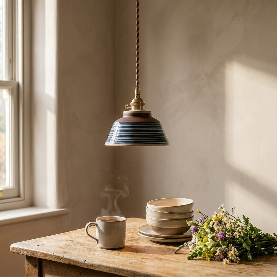 A rustic pendant light with a striped blue and brown ceramic shade hangs over a wooden table in a sunlit room. The table holds a gray ceramic mug, a stack of beige bowls, and a bouquet of wildflowers.