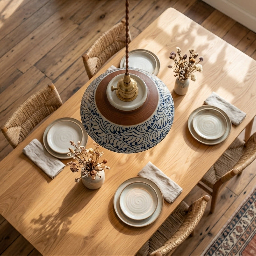 A dining table set in a bright room with a large hanging pendant lamp featuring a blue and white patterned ceramic shade and a brown ceramic center. The table is surrounded by woven chairs and set with plates and dried floral arrangements.